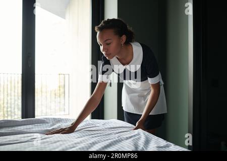 Side view of young housemaid putting clean bedclothes on bed while preparing room of luxurious five star hotel for new guests Stock Photo