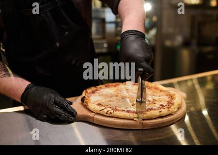 Close-up of young male cook in gloves cutting appetizing pizza on wooden board after baking it while standing by kitchen table Stock Photo