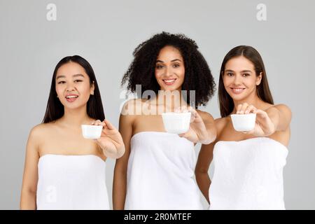 United in hydration. Happy three diverse ladies showing cosmetic face cream jars to camera, sharing cream secrets Stock Photo