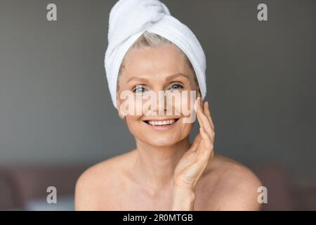 Beautiful senior woman applying face cream and smiling, sitting in bedroom with towel on head after morning shower Stock Photo