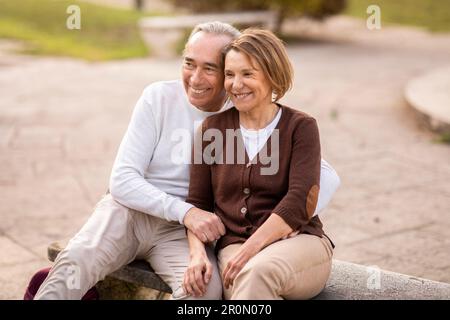 Loving Mature Couple Embracing Sitting On Bench In Park Outside Stock Photo