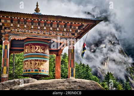 Prayer wheel on a rock in the forest on the ascent to the monastery Taktshang (in the background) or Taktsang or Tigernest, a Buddhist monastery in th Stock Photo