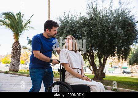 Front view of full body of cheerful husband with disabled wife in wheelchair in casual clothes on tiled street coming from clinic looking at each othe Stock Photo
