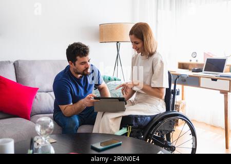 Bearded man sitting on sofa with wife in wheelchair sitting on wheel chair in casual clothes discussing and watching video on tablet Stock Photo