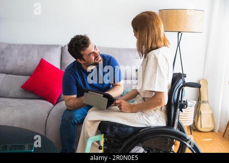 Bearded man sitting on sofa with wife in wheelchair sitting on wheel chair in casual clothes discussing and watching video on tablet Stock Photo