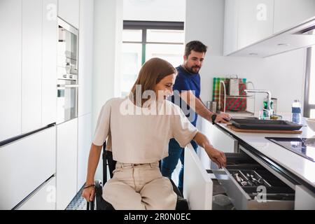 Adorable husband with wife sitting in wheelchair in casual clothes happily discussing while preparing meal together in modern kitchen Stock Photo