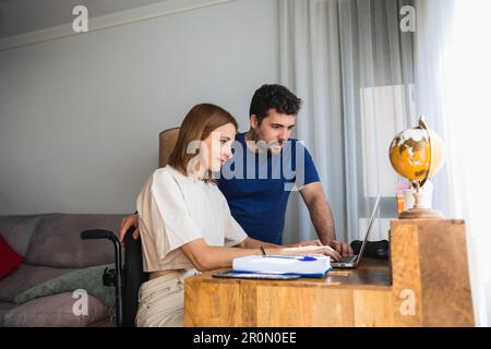 Adorable loving husband assisting wife sitting in wheelchair with office project at home using laptop Stock Photo