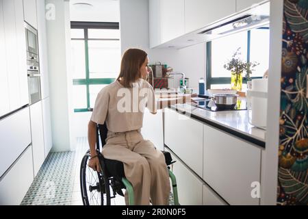 Busy young female in casual clothes sitting on wheelchair by drawer while doing house chores in kitchen in modern apartment Stock Photo