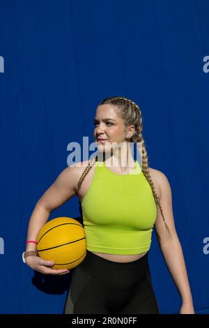 Confident young female athlete in sportswear relaxing on ground and looking away while standing with basket ball against blue wall in daylight Stock Photo