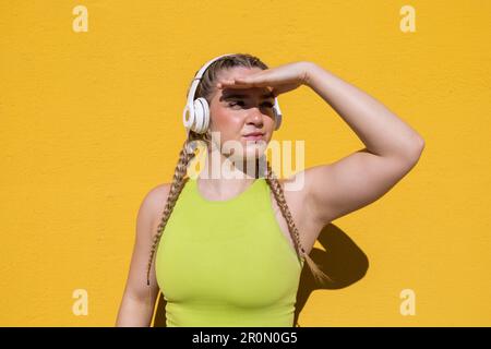 Focused young female in headphones looking away with hand on eyes in sunlight while standing against yellow background and listening to music Stock Photo