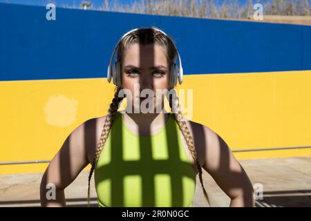Portrait of confident young female in sport wear with braided hair and headphones looking at camera while standing with spread arms against yellow and Stock Photo