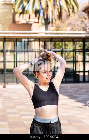 Young fit female in sportswear smiling and looking away while doing ponytail during workout on street Stock Photo