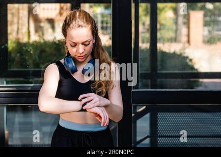Focused young sportswoman wearing wireless headset and activewear setting up smart watch before running during morning workout in city Stock Photo