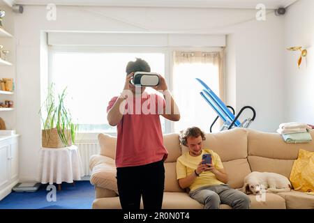 Positive transgender male exploring cyberspace with VR headset while spending time with boyfriend browsing smartphone in living room Stock Photo
