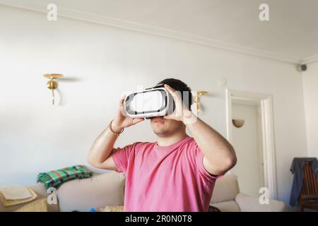 Young male in pink shirt using virtual reality goggles and exploring cyberspace while spending time at home Stock Photo