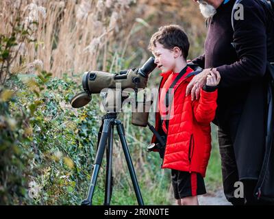 Side view of boy looking through professional photo camera with telephoto lens while standing near crop elderly male photographer in wild nature Stock Photo