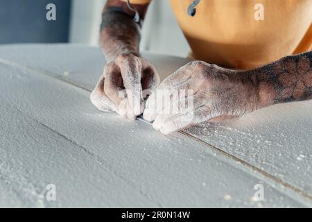 Unrecognizable tattooed male master using special equipment to add stringer on surfboard during work in professional workshop Stock Photo