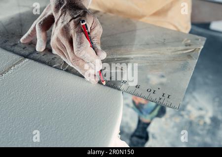 Anonymous tattooed male master in yellow t shirt with respiratory mask on neck using special measuring tool and pencil for making marks on surfboard d Stock Photo