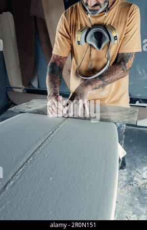 Anonymous tattooed male master in yellow t shirt with respiratory mask on neck using special measuring tool and pencil for making marks on surfboard d Stock Photo