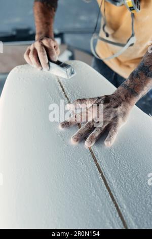 Unrecognizable tattooed male master polishing and sanding surfboard while using special instrument in light workshop during work process Stock Photo