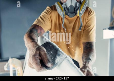 Cropped unrecognizable adult bearded male master with respirator on mask polishing border of surfboard with sandpaper while working in workshop Stock Photo