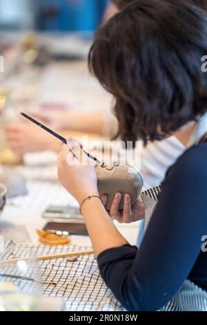 Cropped unrecognizable concentrated woman in apron using brush to paint while creating handmade clay on table with fabric during craft Stock Photo