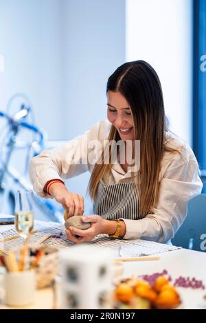 Cheerful young female artisan in apron making handmade clay bowl and smiling while working at workbench in professional pottery studio Stock Photo
