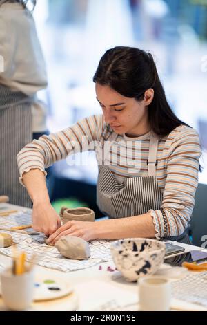 Young female in apron standing at table with various tools and forming handmade clay vase during pottery Stock Photo