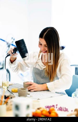 Positive young female artist in apron smiling and using dryer for clay cup while making earthenware during pottery process in studio Stock Photo