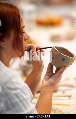 Side view of serious young female artist in white shirt applying colorful dots on clay cup with paintbrush while working in professional pottery studi Stock Photo
