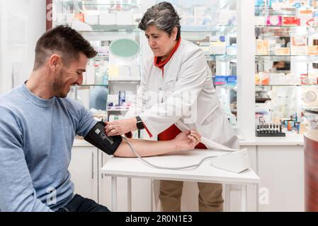 Concentrated female pharmacist in white robe using sphygmomanometer while measuring blood pressure male customer in pharmacy Stock Photo