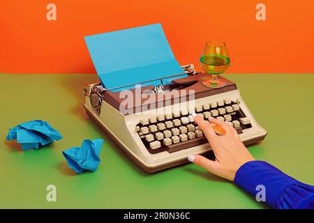 Crop anonymous person at table with glass of wine typing on old typewriter with blue carton paper over green two colored background Stock Photo
