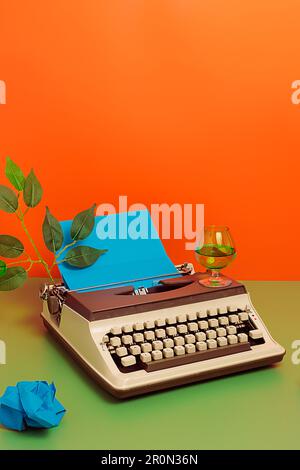 High angle of vintage typewriter with green leaves and crumpled blue papers with glass of wine over green and orange background in studio Stock Photo