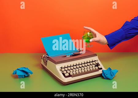 Crop anonymous person in blue clothes with glass of wine and old typewriter with blue paper on set and crumpled paper over two color background in stu Stock Photo