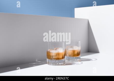 Front view of translucent glasses filled with cold refreshing scotch whiskey with ice cubes placed on white surface against white wall Stock Photo
