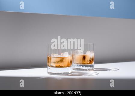 Translucent glasses filled with cold refreshing scotch whiskey with ice cubes placed on white surface against white wall Stock Photo