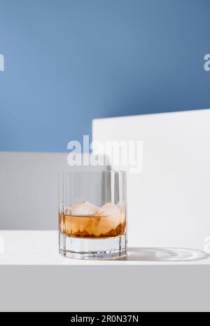 Close up of transparent glass of whiskey with ice cubes placed on white surface against white walls Stock Photo