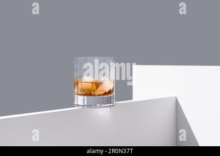 Transparent glass of whiskey on rocks with ice cubes placed on edge of white wooden table isolated on gray background Stock Photo