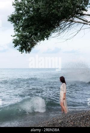 Side view of tranquil young brunette in wet shirt walking along a clear blue sea and looking down against cloudy sky during summer vacation Stock Photo