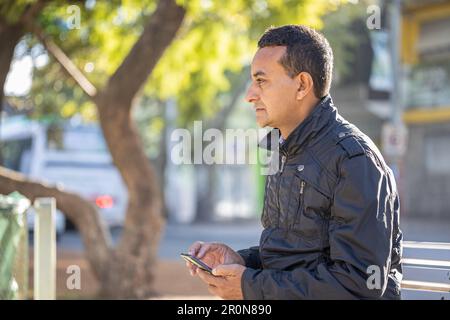 Young latin man sitting on a square bench using his mobile phone. Stock Photo