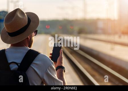 Traveler man wearing straw hat using has mobile phone while standing on railway platform and waiting train. Stock Photo