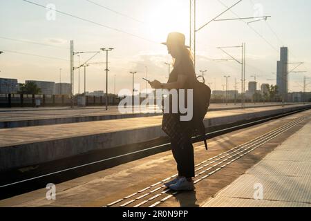 Tourist woman with backpack standing on railway station platform and using phone at sunset. Stock Photo