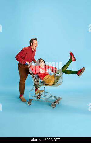 Portrait with couple, man and pretty woman having a shopping cart race over blue color studio background. Adventure time Stock Photo