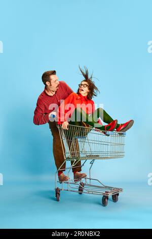 Portrait with funny couple, handsome man and excited woman riding in shopping cart over blue studio background. Black Friday sales Stock Photo