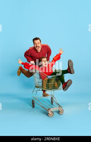 Portrait with couple, man and pretty woman having a shopping cart race over blue color studio background. Adventure time Stock Photo