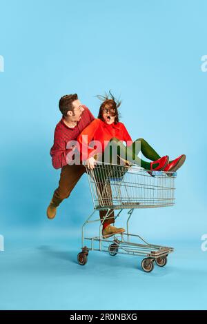 Portrait with funny couple, man and attractive woman sitting at shopping cart and riding on blue background. Crazy family Stock Photo