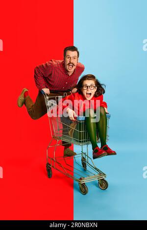 Portrait with funny couple, man and attractive woman sitting at shopping cart and racing on blue background. Crazy family Stock Photo