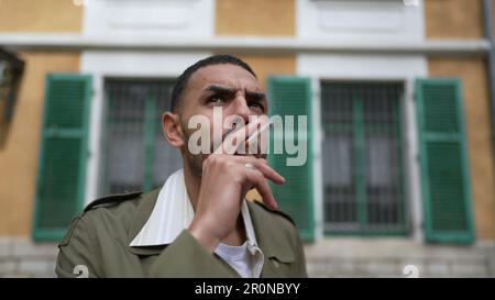 One pensive serious young Middle Eastern man smoking a cigarette outside with thoughtful expression. Tracking shot closeup face in motion while broodi Stock Photo
