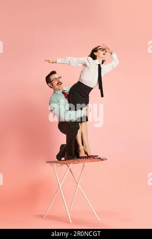 Portrait with funny couple, handsome man and happy woman riding on ironing board like surfing over pink background. Black Friday sales Stock Photo