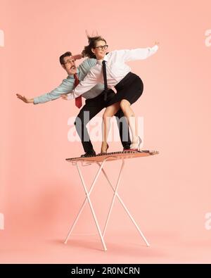 Portrait with funny couple, handsome man and excited woman riding on ironing board like surfing over pink background. Black Friday sales Stock Photo